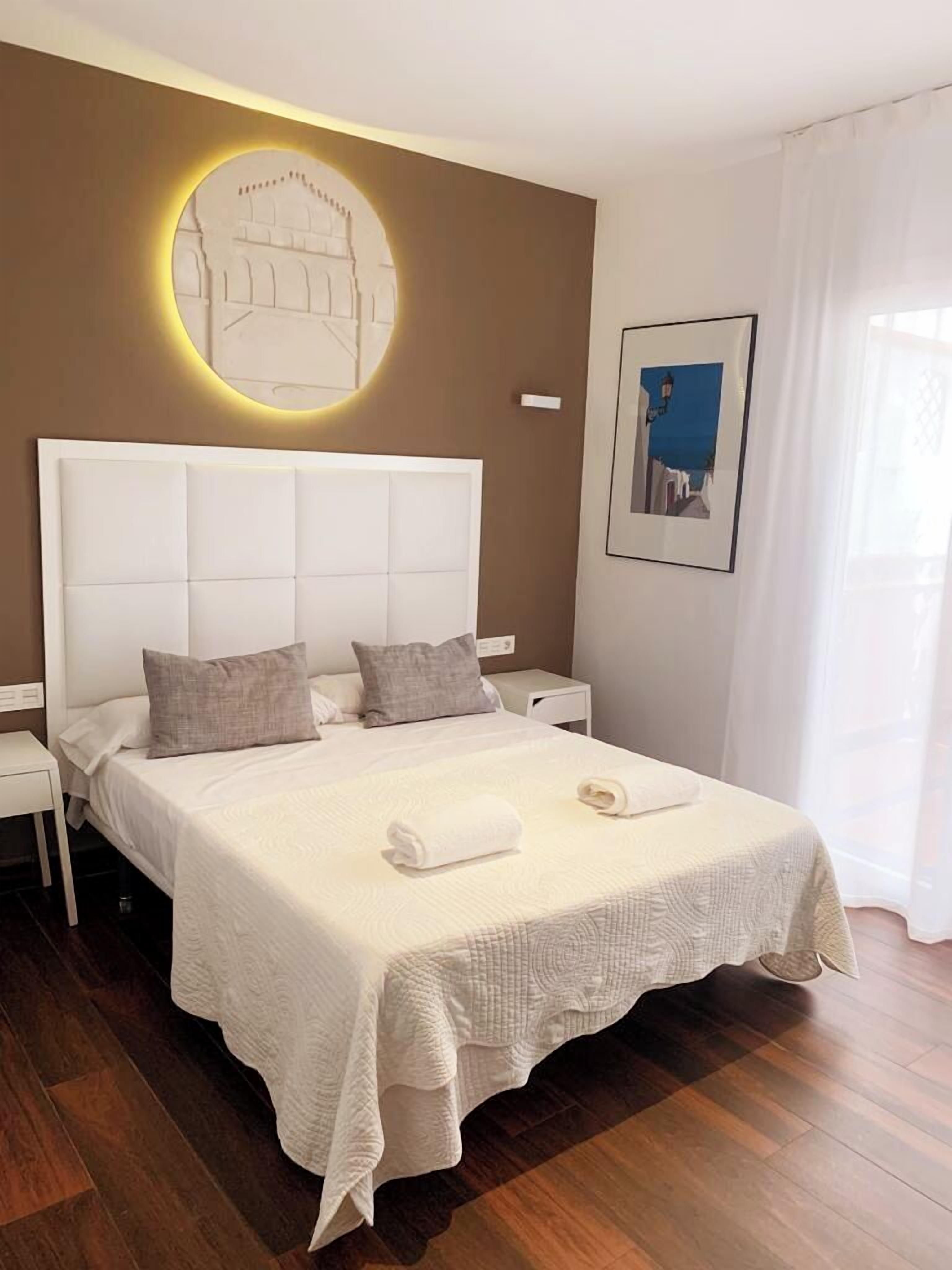 A neatly arranged bedroom with a large bed, a bedside table, a wall-mounted light fixture, and a framed picture.