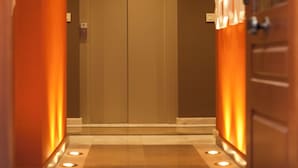 Un pasillo de hotel con una puerta doble, una lámpara fijada a la pared y un adorno mural con motivos geométricos.