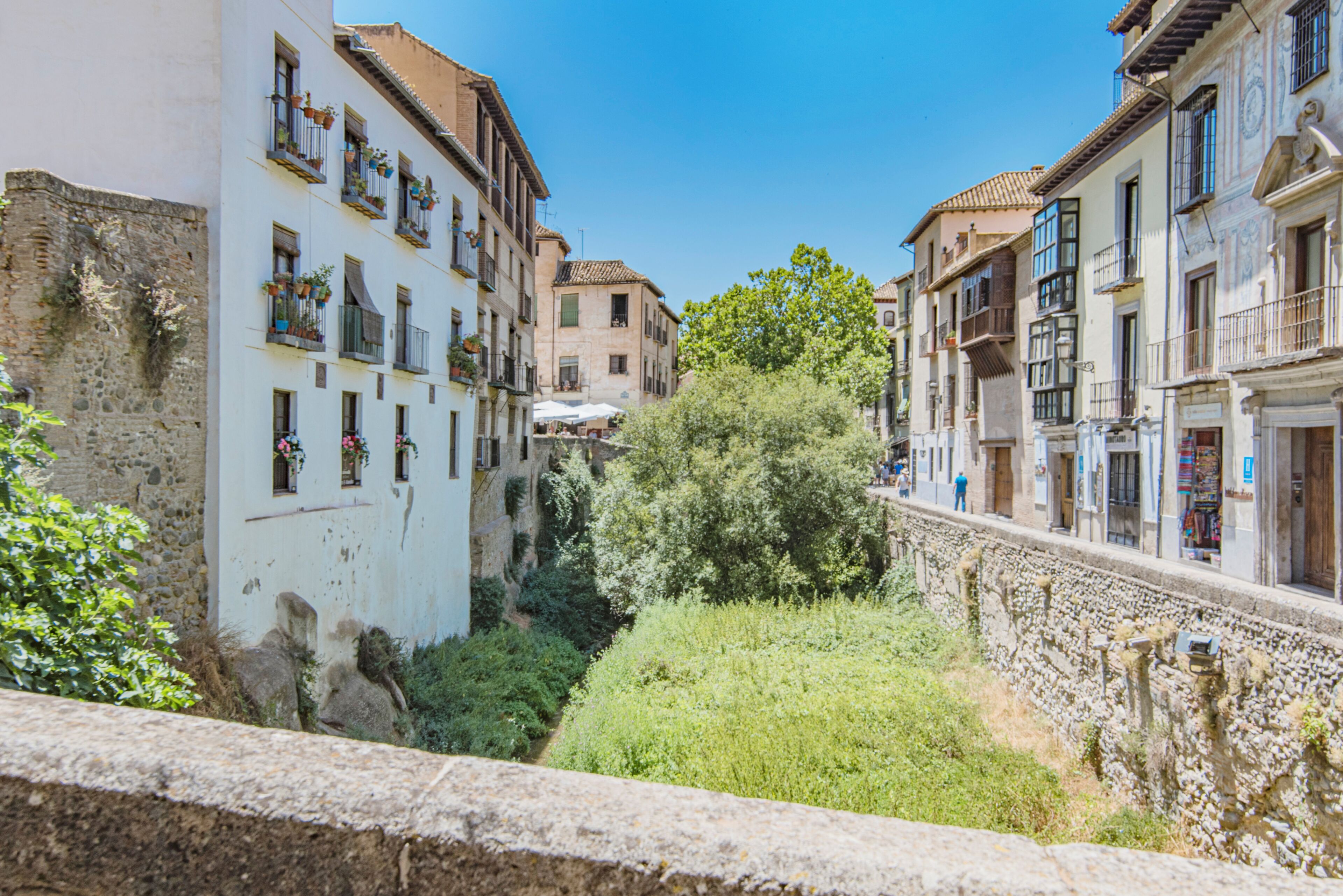 Una calle estrecha bordeada por edificios con balcones, una pared de piedra y vegetación.