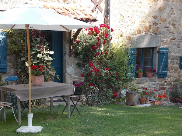 A rustic outdoor seating area with a table and chairs under a white umbrella, surrounded by potted plants and a stone building with blue shutters. - La Rosière 2. A heavenly place in the midst of nature, peace and quiet. (Auzelles)