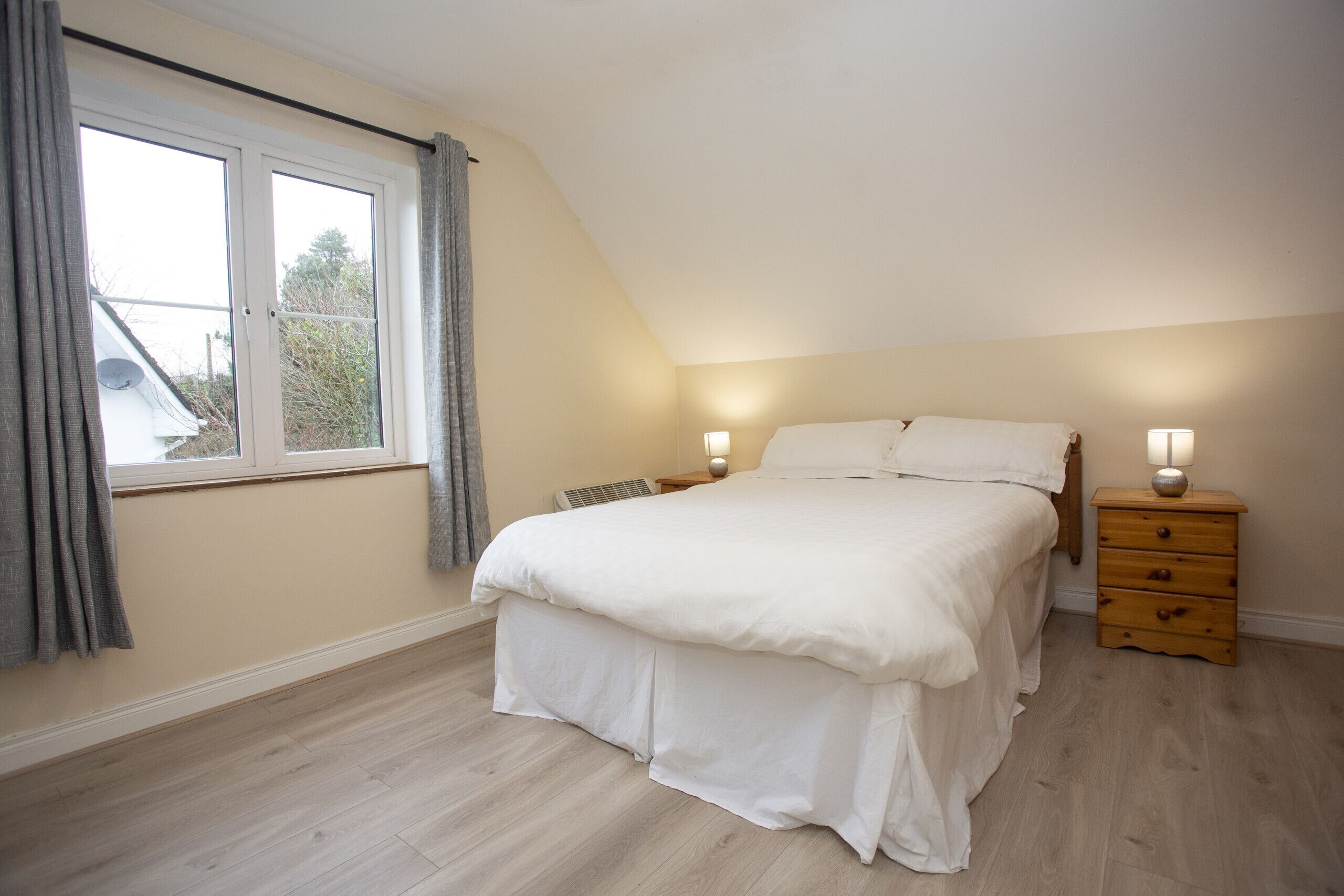 Ein Schlafzimmer mit einem großen Bett, zwei Nachttischen mit Lampen, einem Fenster mit Vorhängen und einer geneigten Decke.