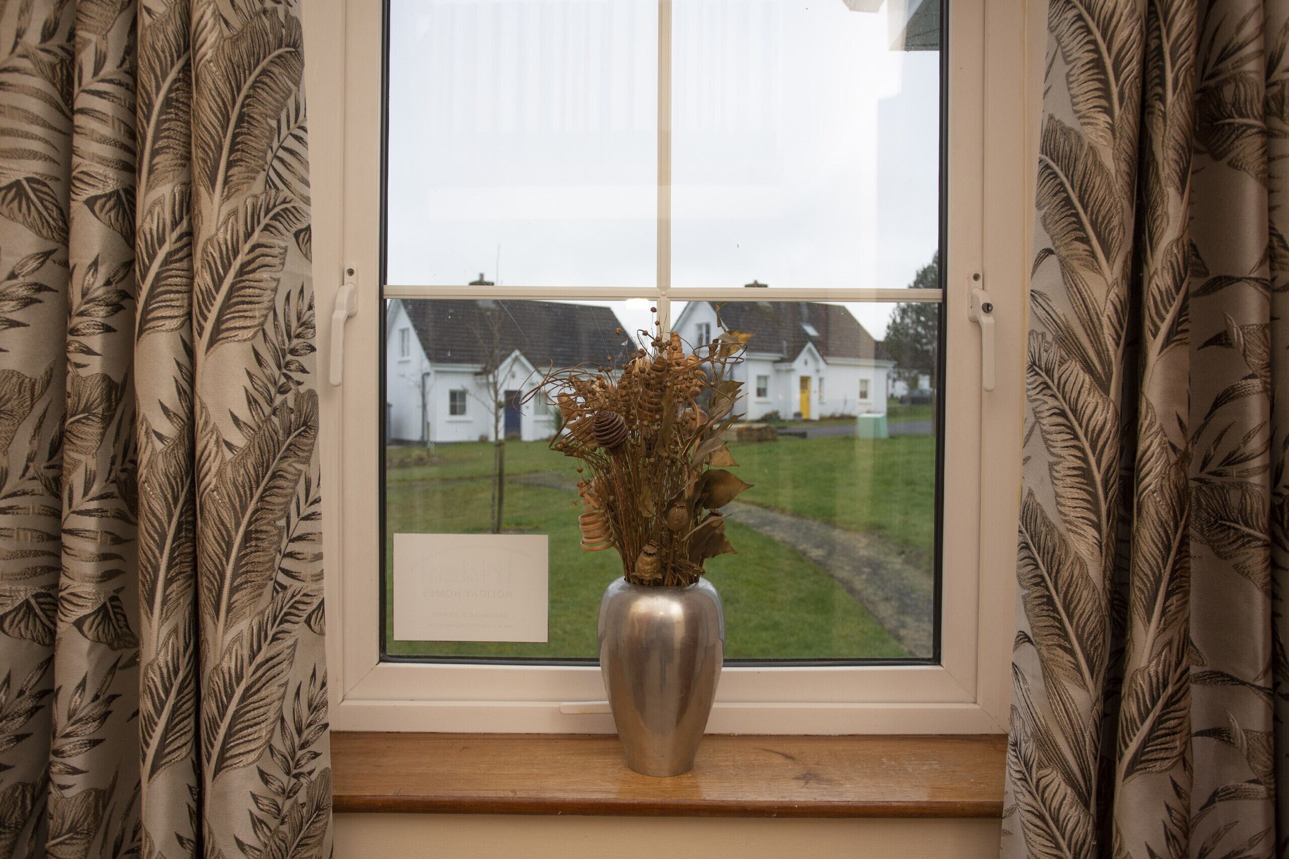A window with a view of houses and a vase with dried flowers on a windowsill.