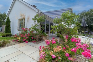 Una casa blanca con un porche cubierto, rodeada de un jardín con diversas plantas en flor.
