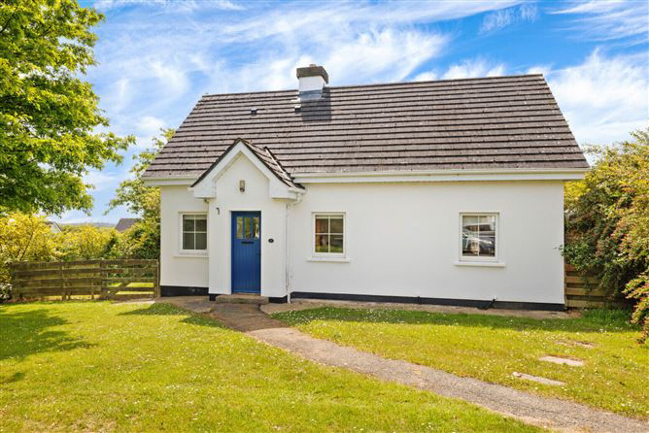 A single-story white house with a blue door, a chimney, and a small front yard with a wooden fence.