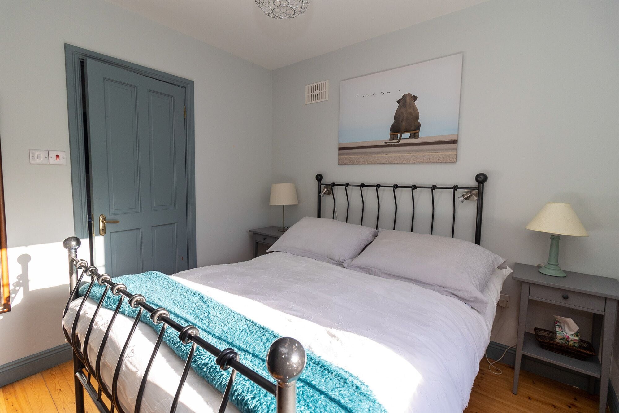 A bedroom with a bed, two bedside tables, a nightstand, a blue door, and a painting on the wall.
