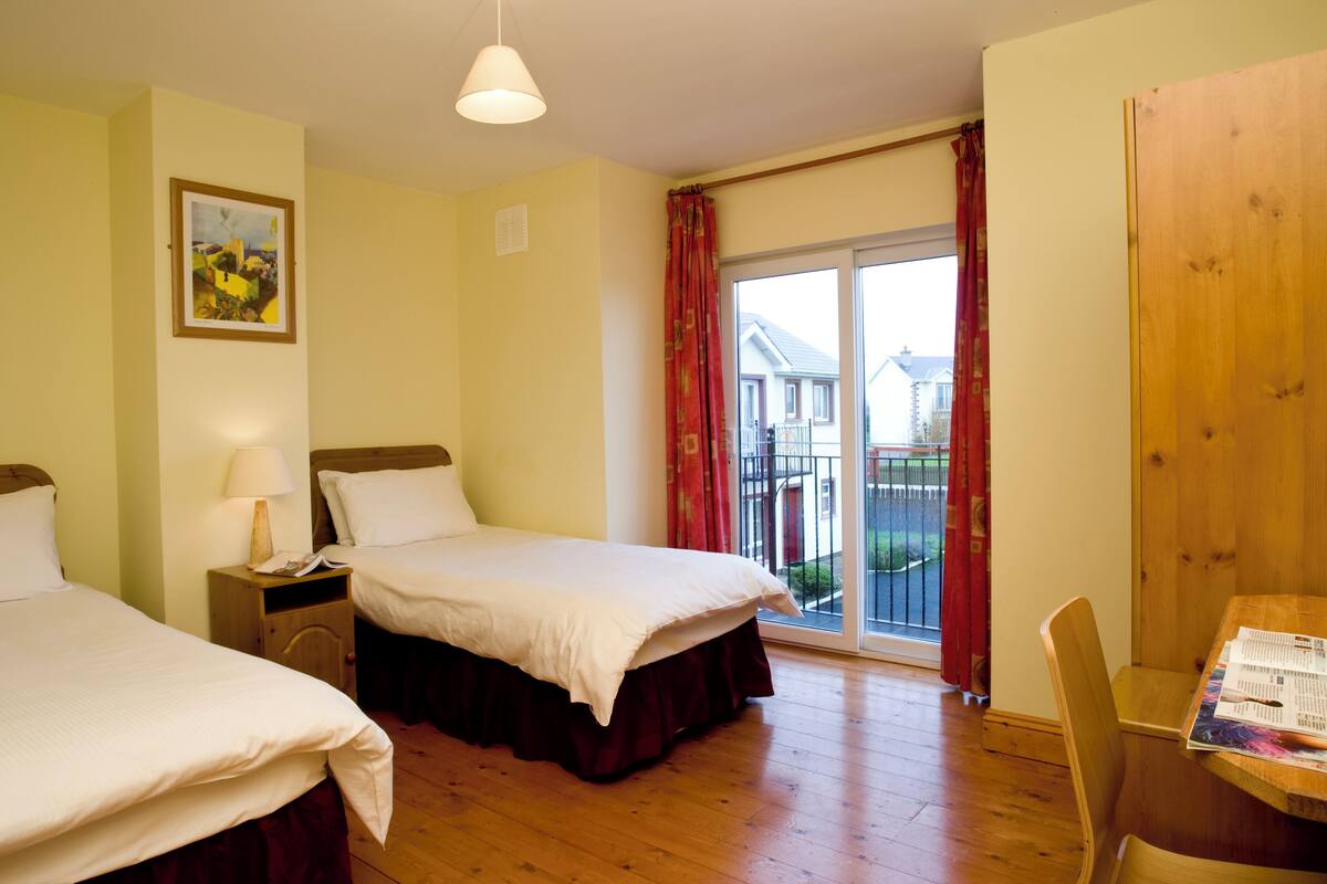 A hotel room with two beds, a wooden table, a chair, a window with a view of houses, and a door.