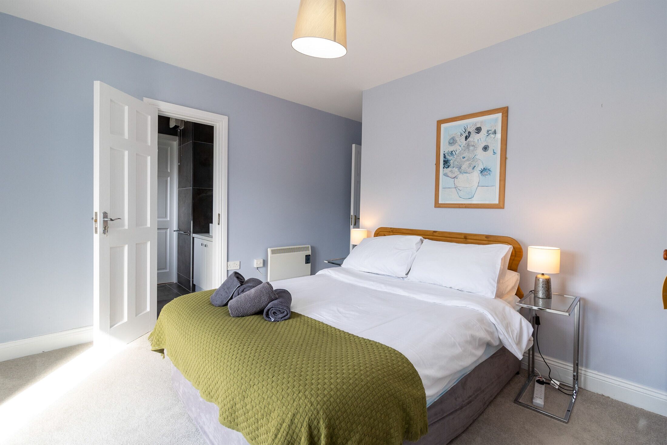 A neatly made bed with a green blanket, a bedside lamp, and a framed artwork on the wall.