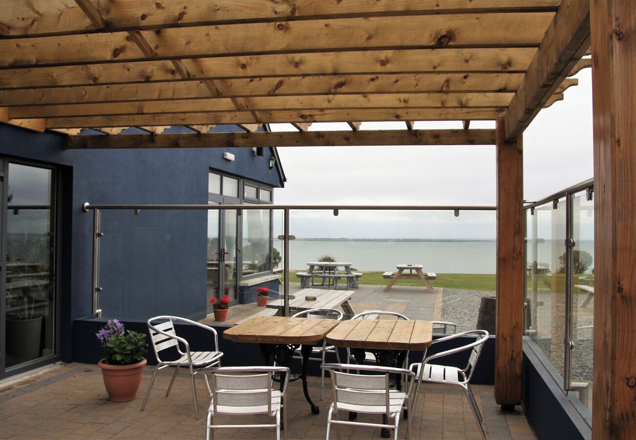 Un patio cubierto con mesas y sillas de madera, vista al mar y un edificio con grandes ventanales.
