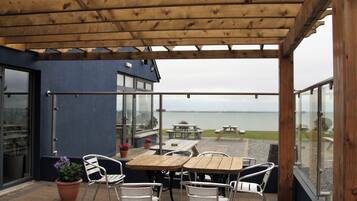 A covered patio with wooden tables and chairs, a view of the sea, and a building with large windows.