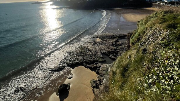 Un paisaje costero con cielo despejado, una playa de arena y afloramientos rocosos.