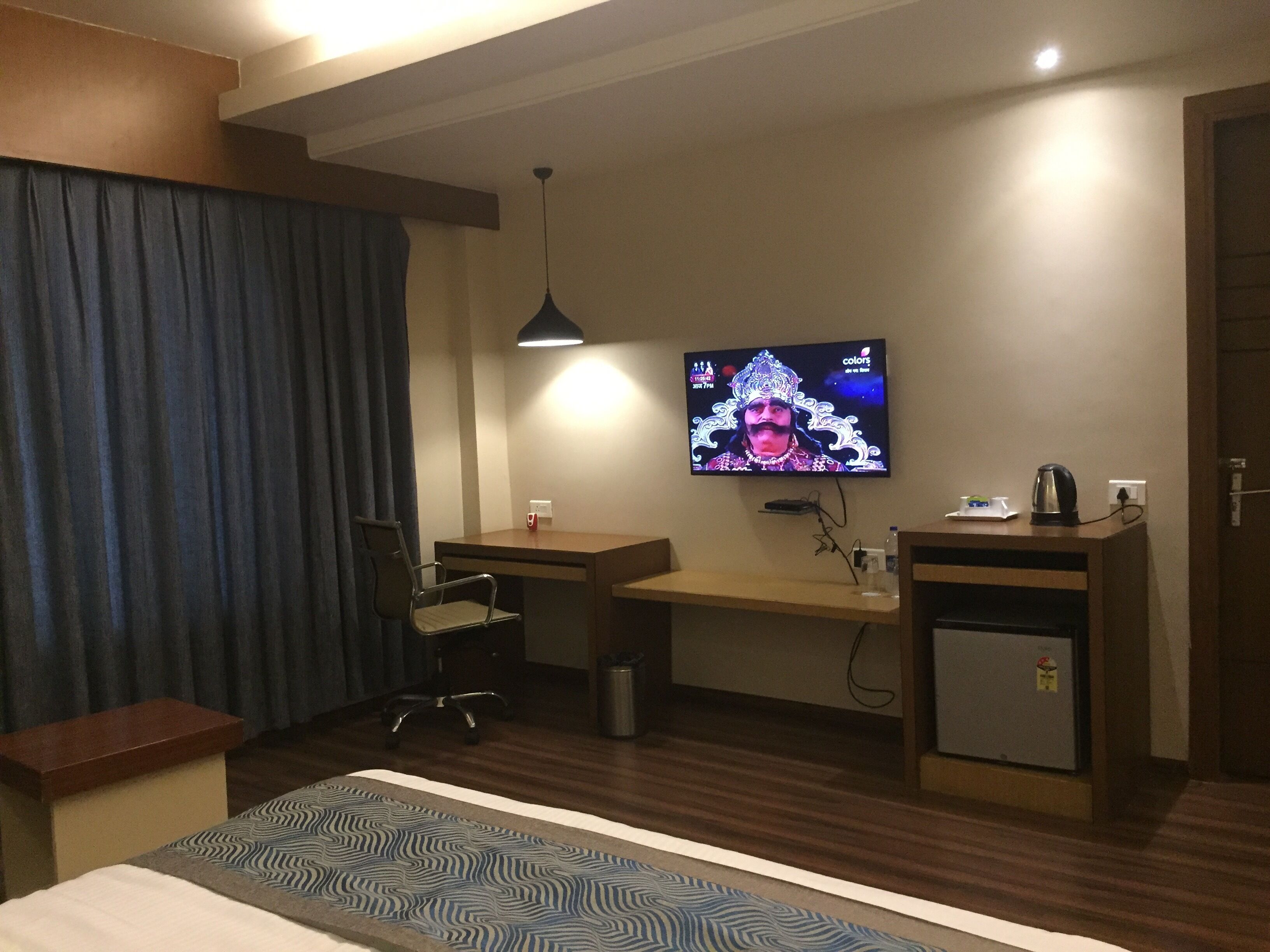 A hotel room with a bed, a desk, a chair, a TV mounted on the wall, and a small cabinet.