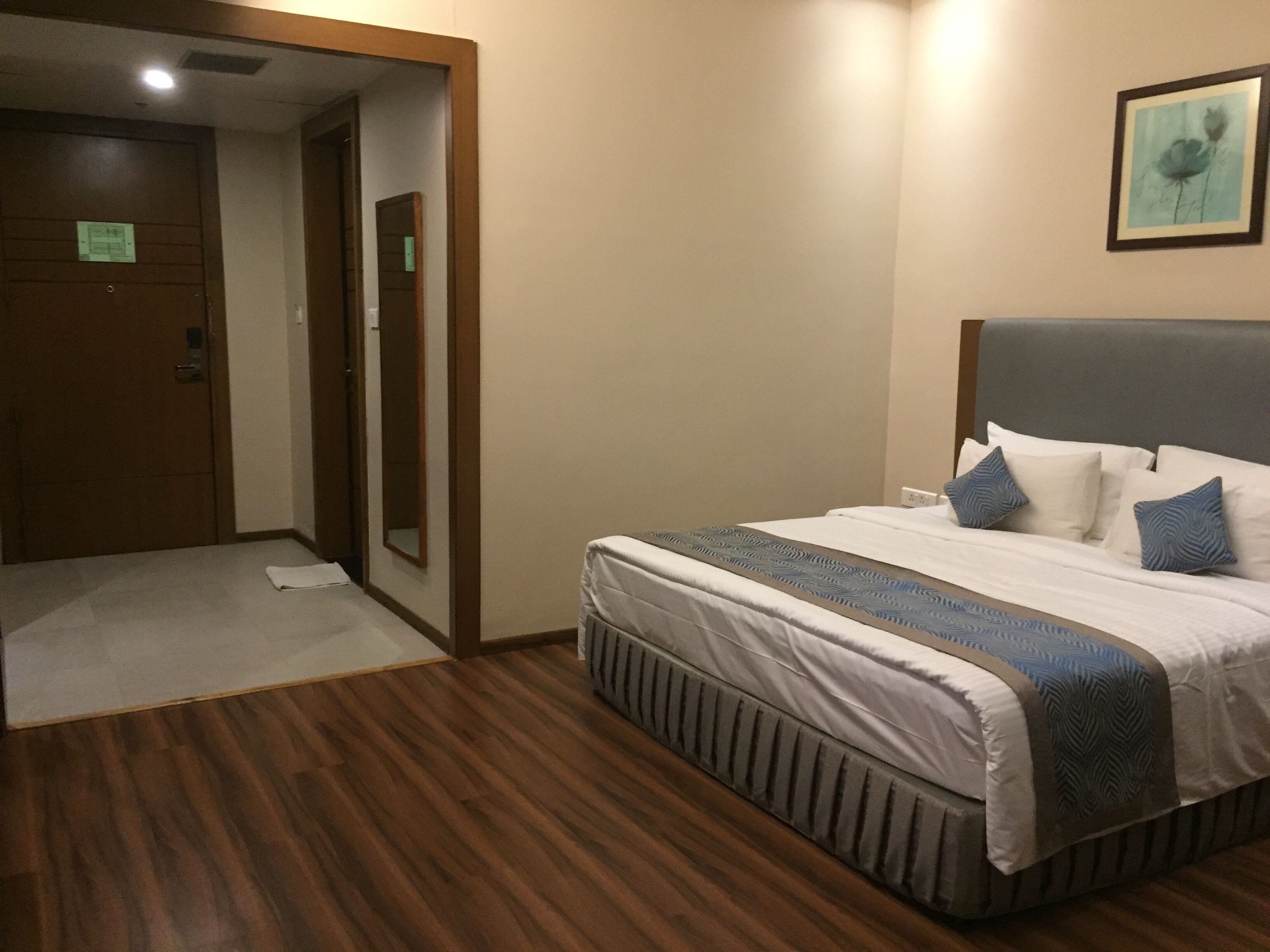 A hotel room with a wooden floor, a bed with white bedding and blue pillows, a large mirror, and a framed picture on the wall.