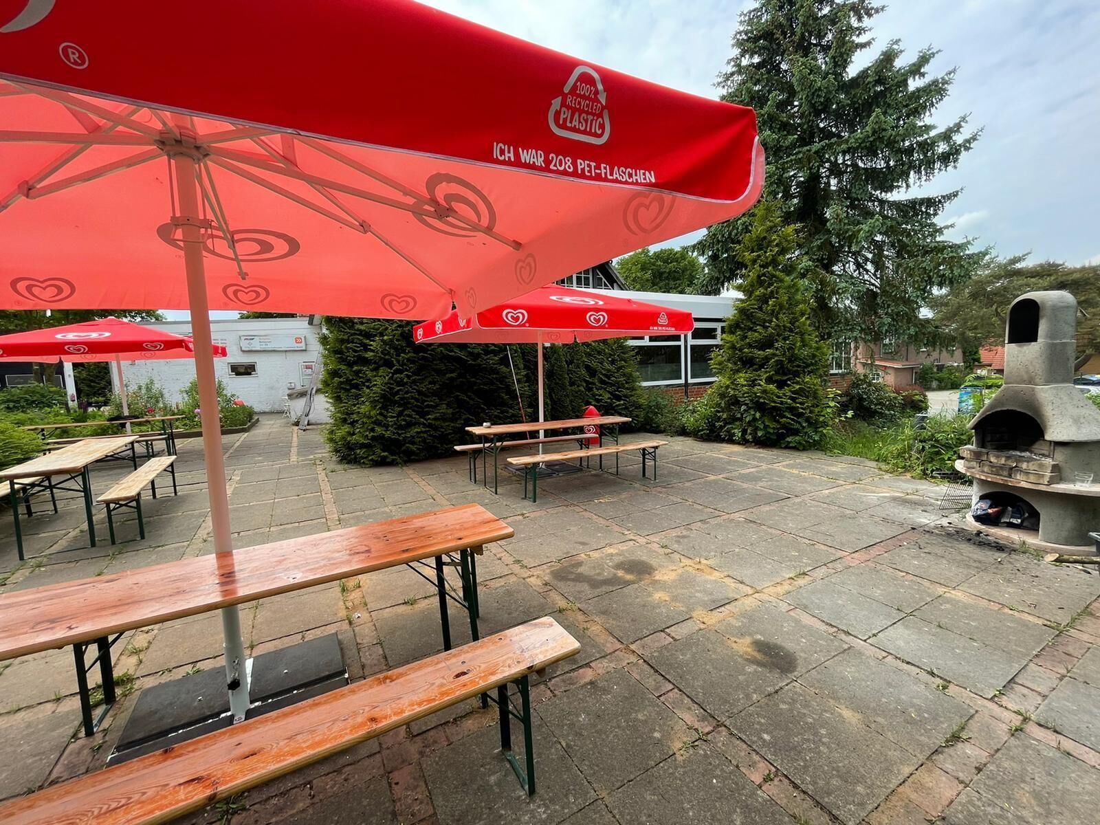 An outdoor patio area with red umbrellas, wooden benches, and a stone oven.