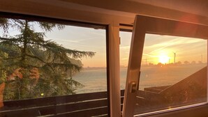 A view from a window with a wooden railing, overlooking a body of water and a forested area with wind turbines in the distance. - K357 - Monteurhotel Ostsee Lounge (Ratekau)