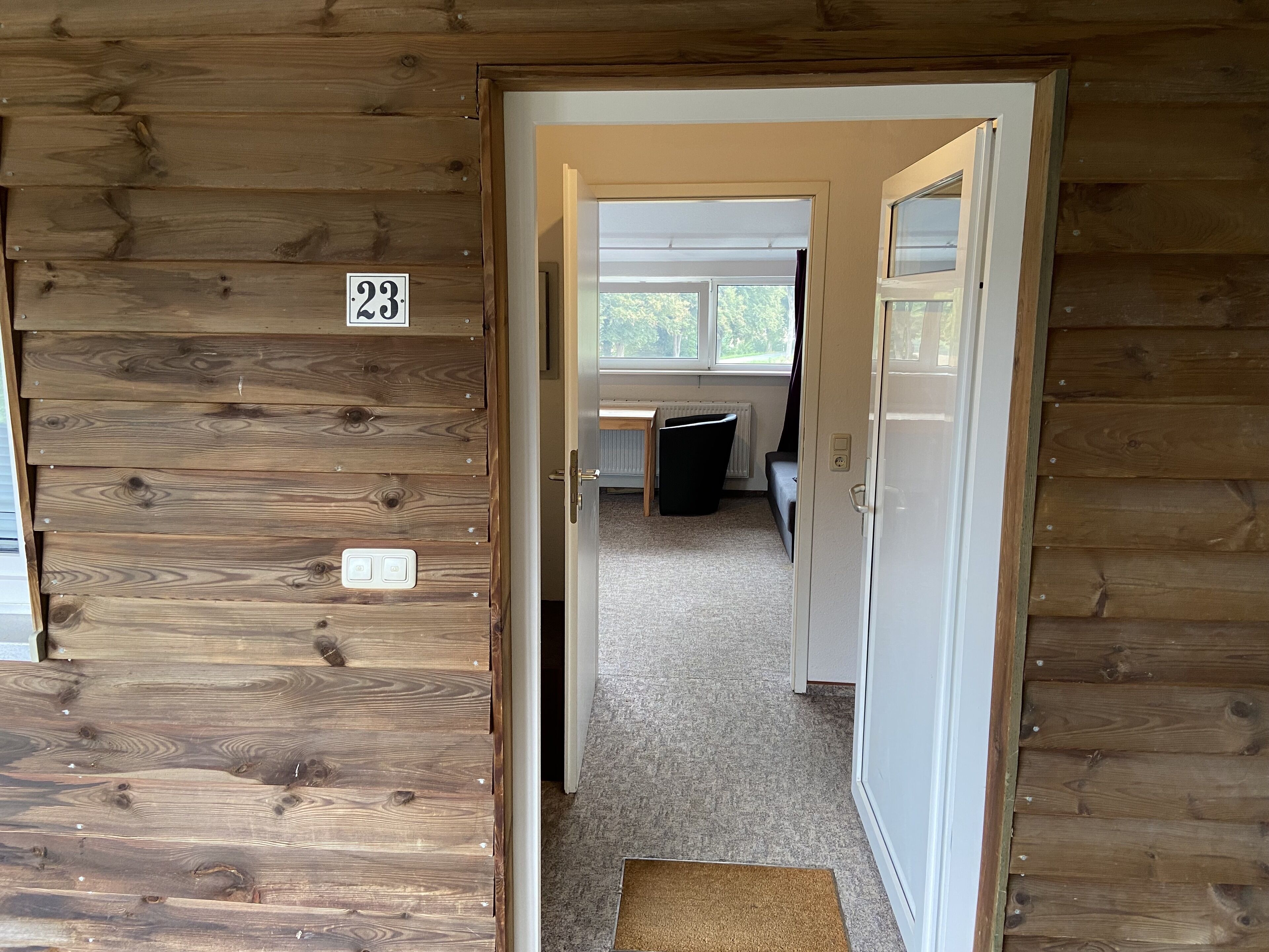 A wooden interior with a door marked '23' leading to a room with a window, a radiator, and a black chair.