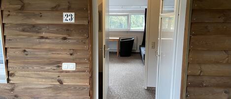 A wooden interior with a door marked '23' leading to a room with a window, a radiator, and a black chair.