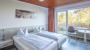 A double bed with white linens and pillows, a bedside table with a lamp, a window with curtains, and a view of trees outside. - K357 - Monteurhotel Ostsee Lounge (Ratekau)