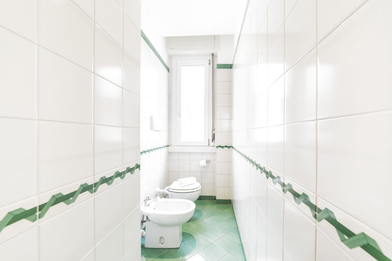 A narrow bathroom with white tiles and green accents, a toilet, bidet, and a sink.