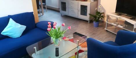 A modern living room with a blue sofa, a glass coffee table, and a colorful rug. There is a kitchen area in the background with a microwave and a coffee machine.