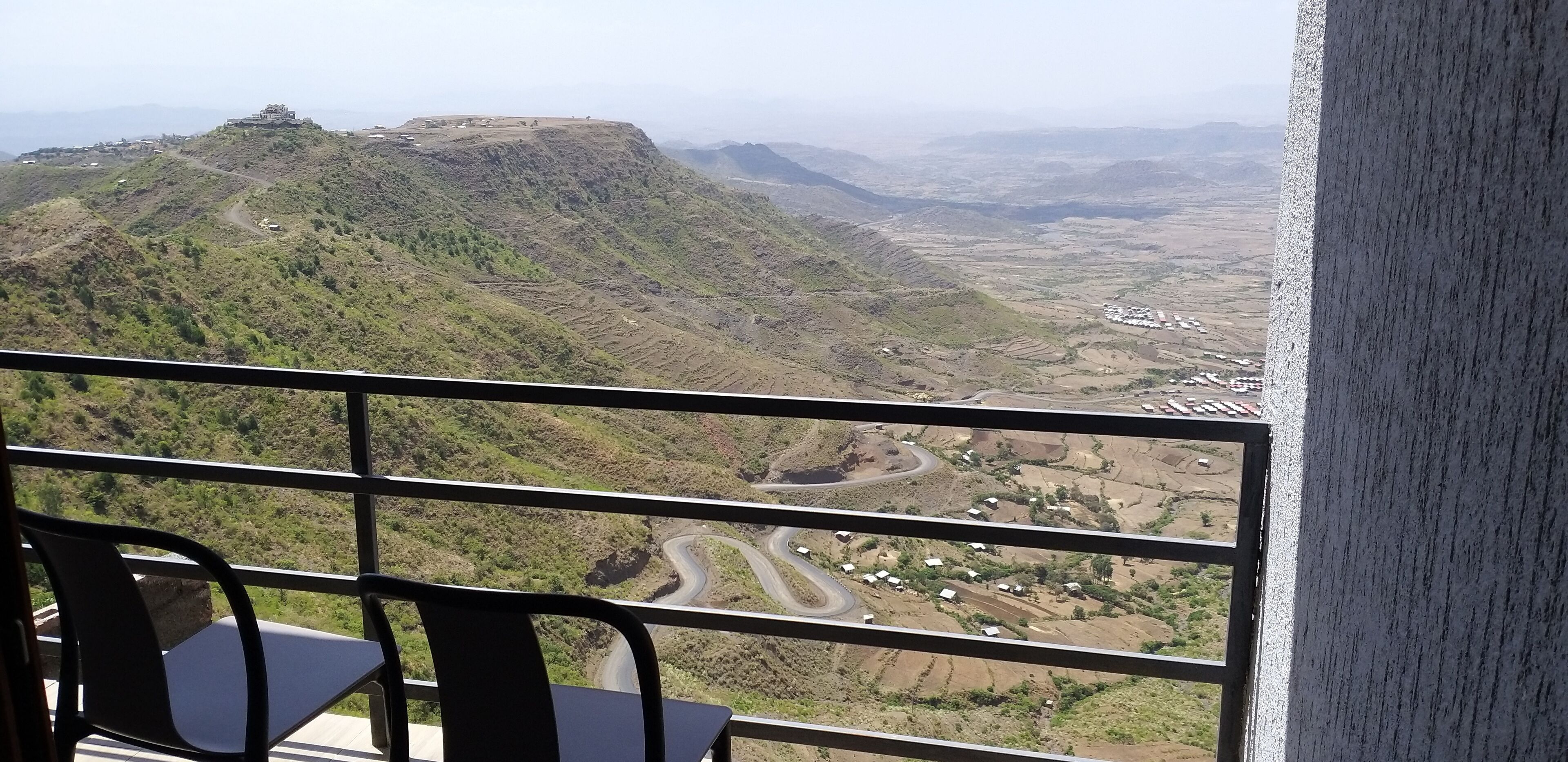 Vista desde un balcón sobre un valle con un camino serpenteante, casas dispersas y una colina destacada.