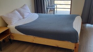 Una cama de madera con una manta gris, una mesita de noche de madera y una silla junto a la ventana.
