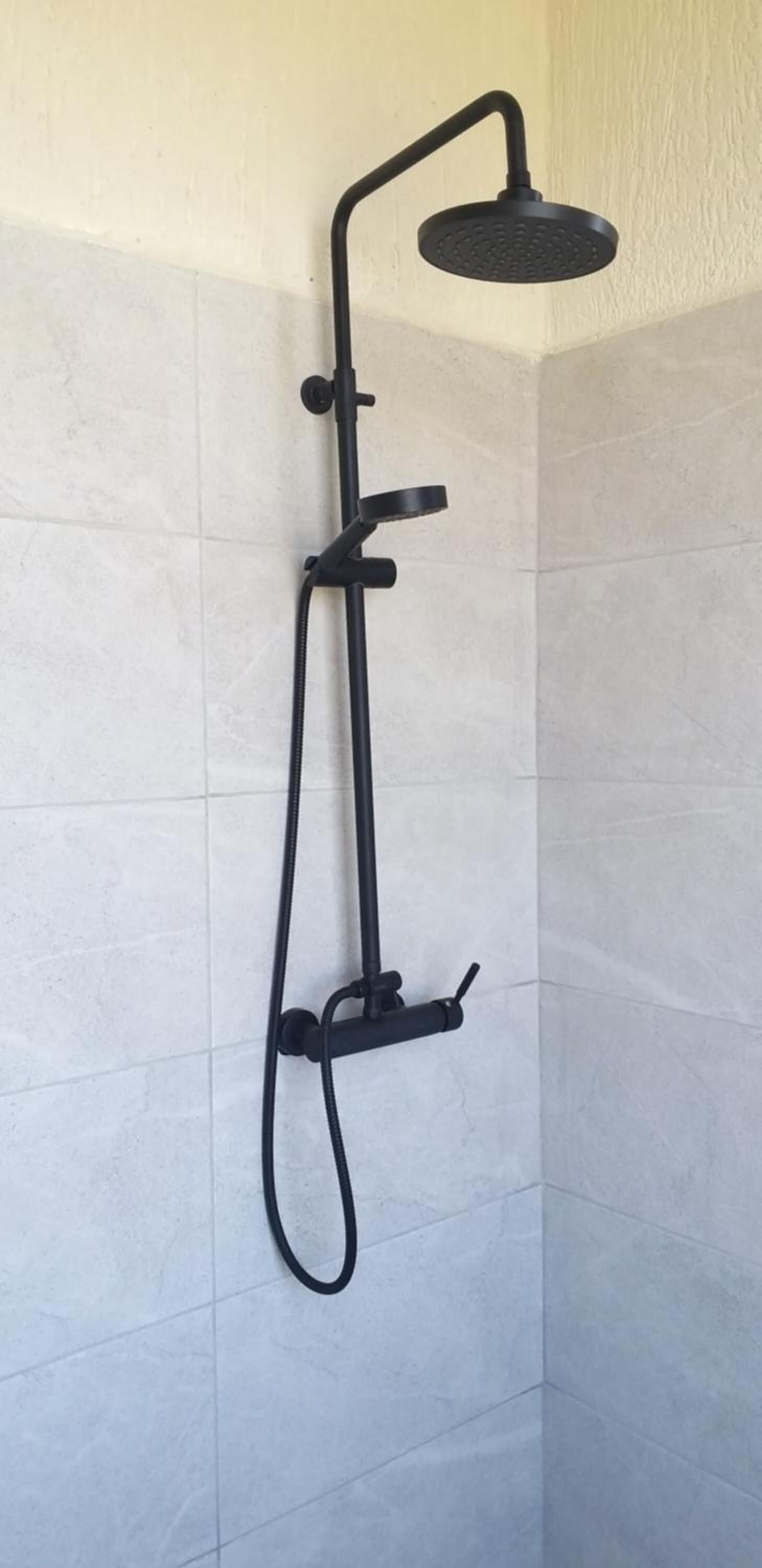 Un cabezal de ducha negro independiente con un rociador de mano, instalado en una pared de azulejos.