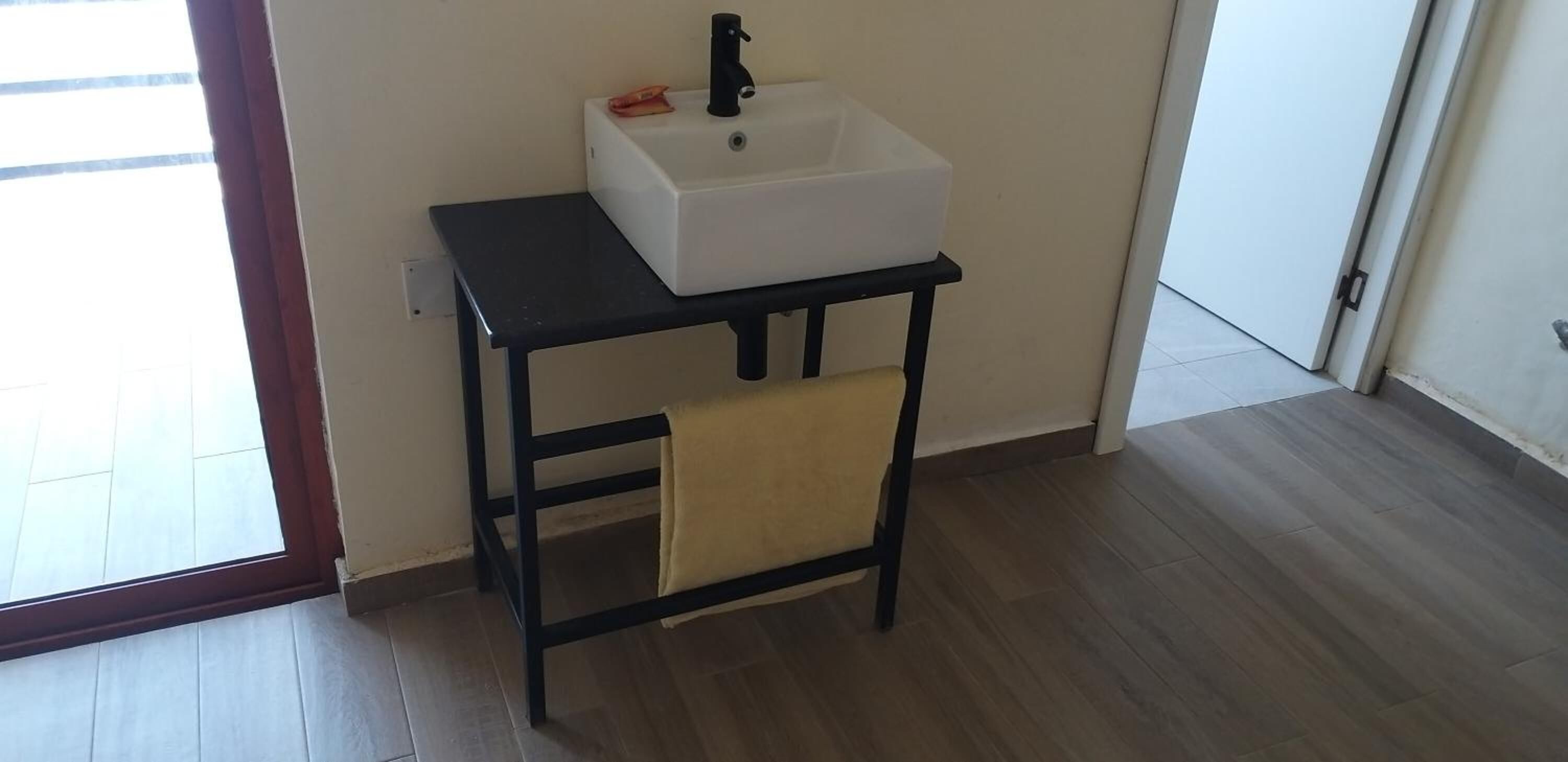 A small bathroom with a white sink on a black table, a towel hanging on the table, and a wooden floor.