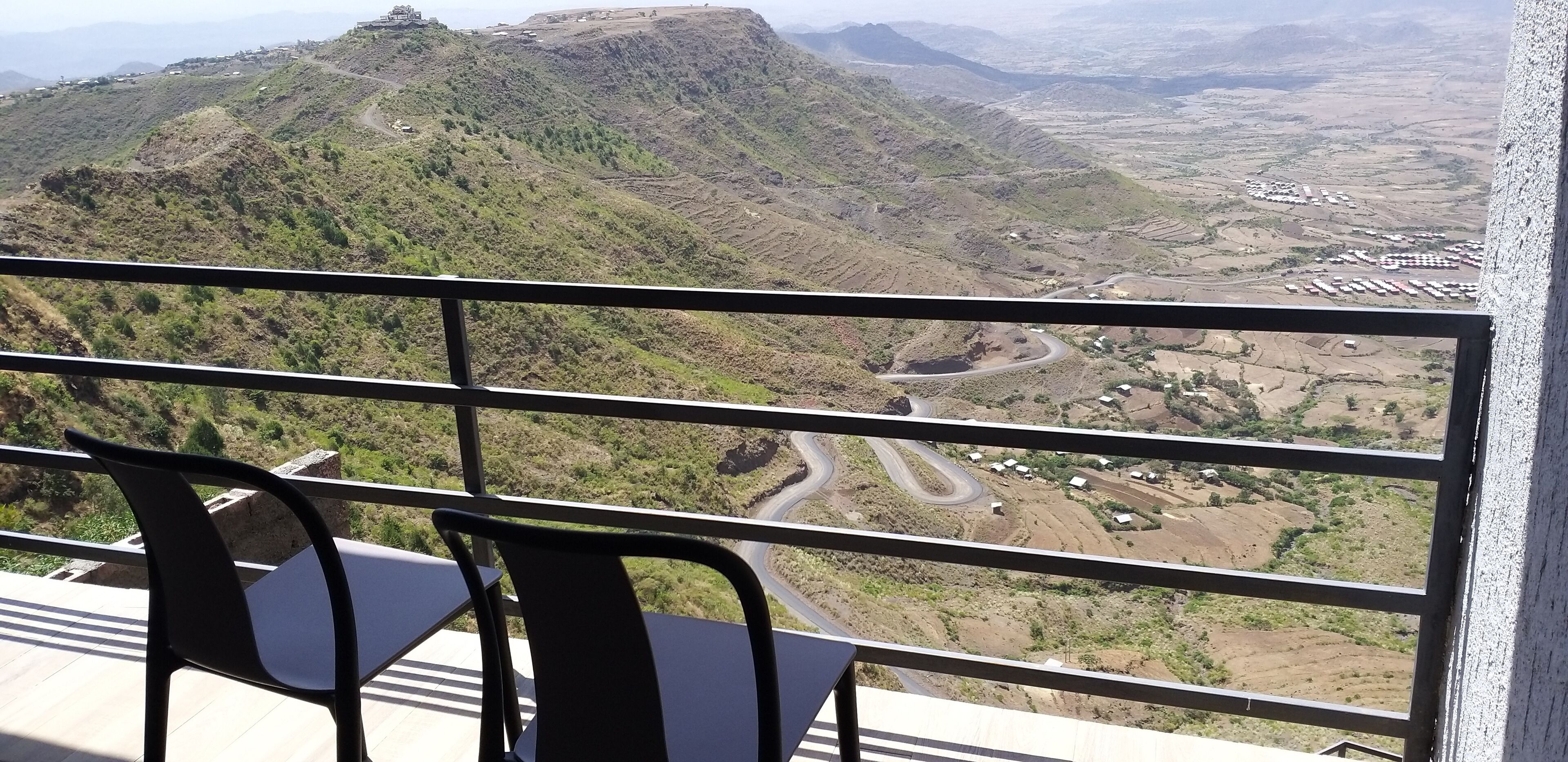 A balcony with a view of a mountainous landscape, including a winding road and scattered buildings.