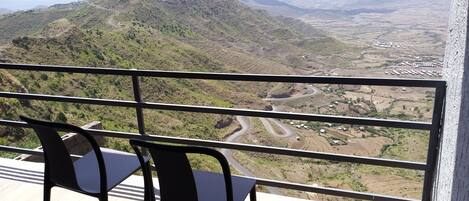 A balcony with a view of a mountainous landscape, including a winding road and scattered buildings.