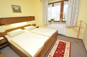 A double bed with a wooden headboard, a nightstand, a bedside table, a window with floral curtains, a radiator, and a red floral rug. - Appartementhaus Alpenrose (Wallgau)