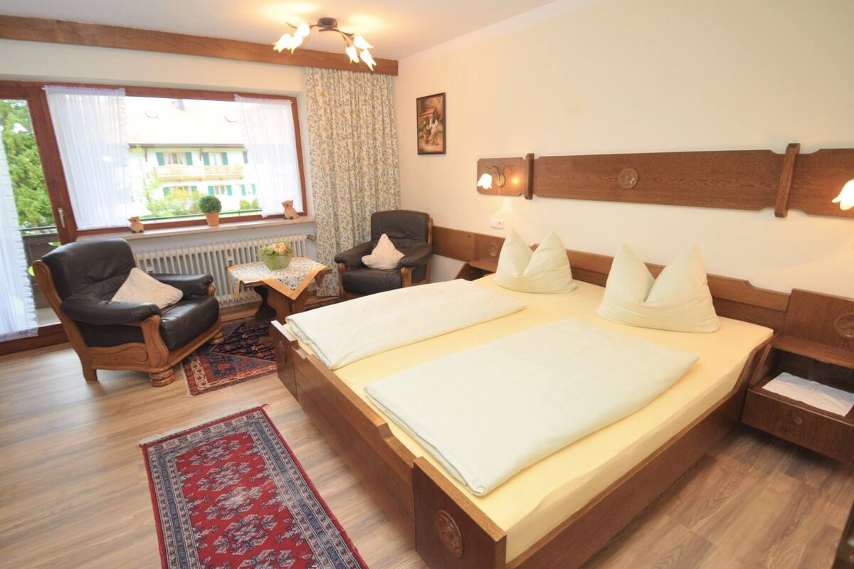 A hotel room with a double bed, two chairs, a small table with a vase, and a view of a building through the window.