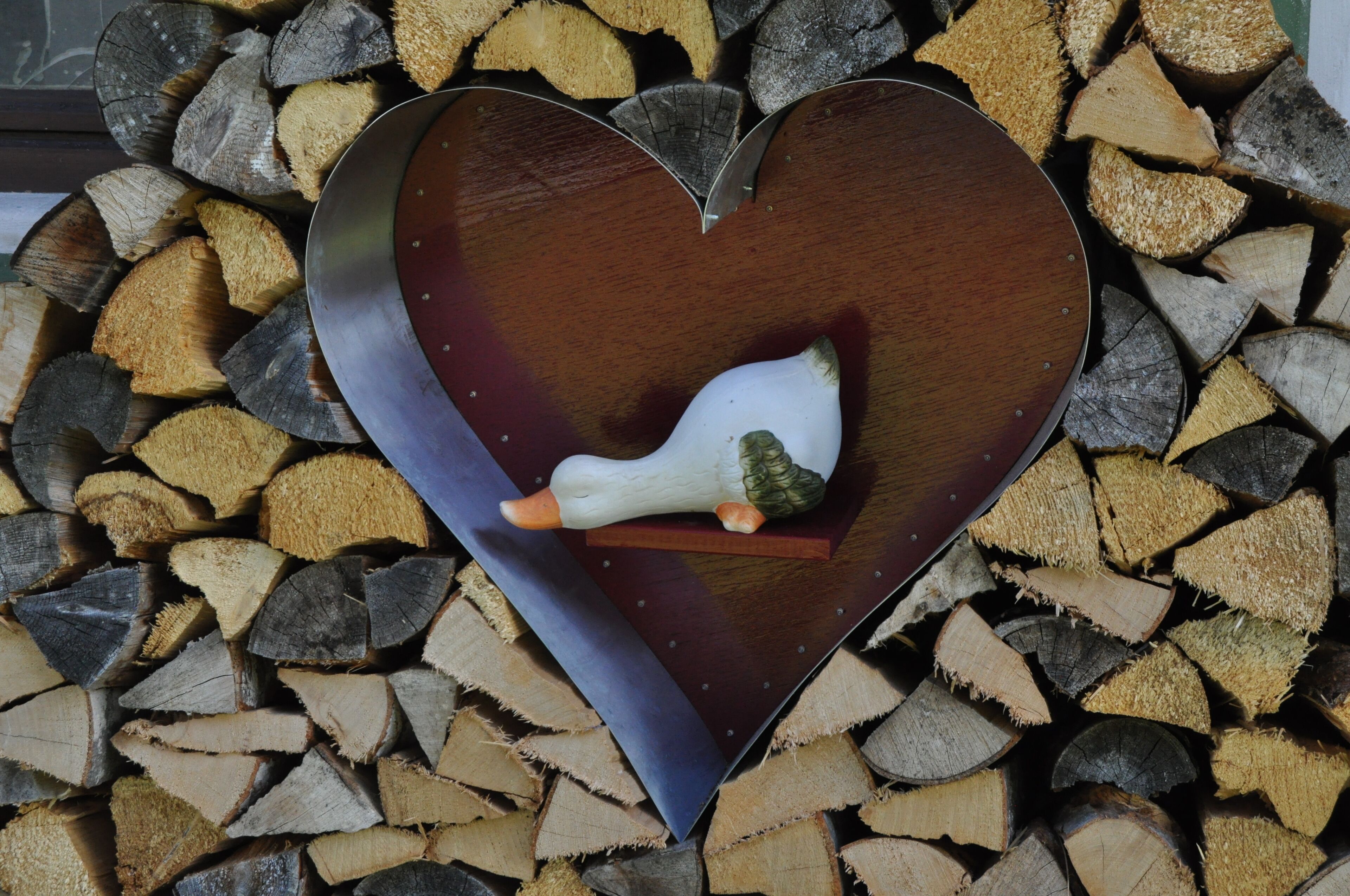 A heart-shaped metal object with a duck figurine on a wooden base, surrounded by stacked logs.