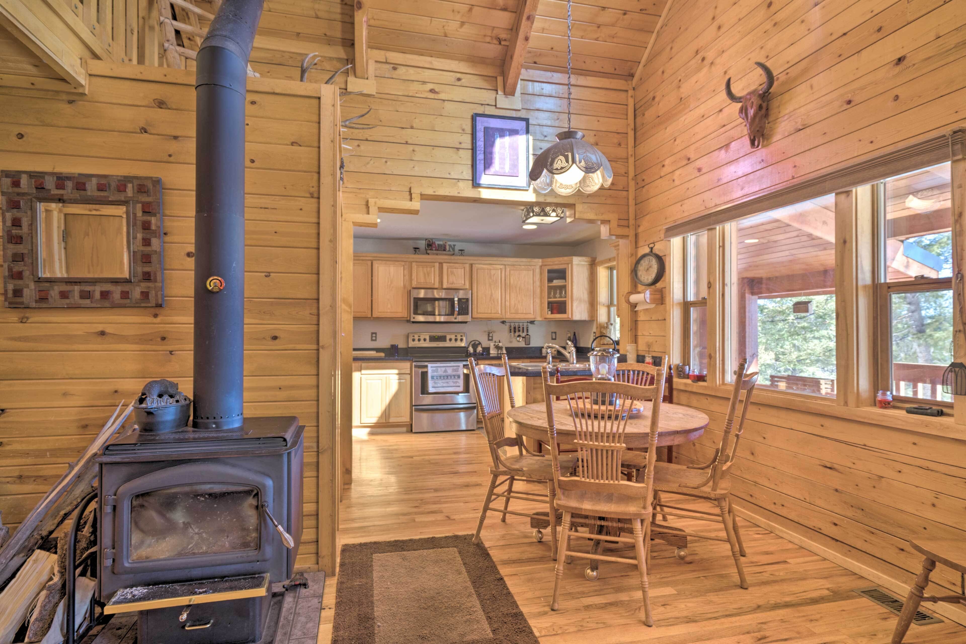 Ein Holzblockhaus-Innenraum mit zentralem Holzofen, einem Essbereich mit Stühlen und einer Küche mit modernen Geräten.