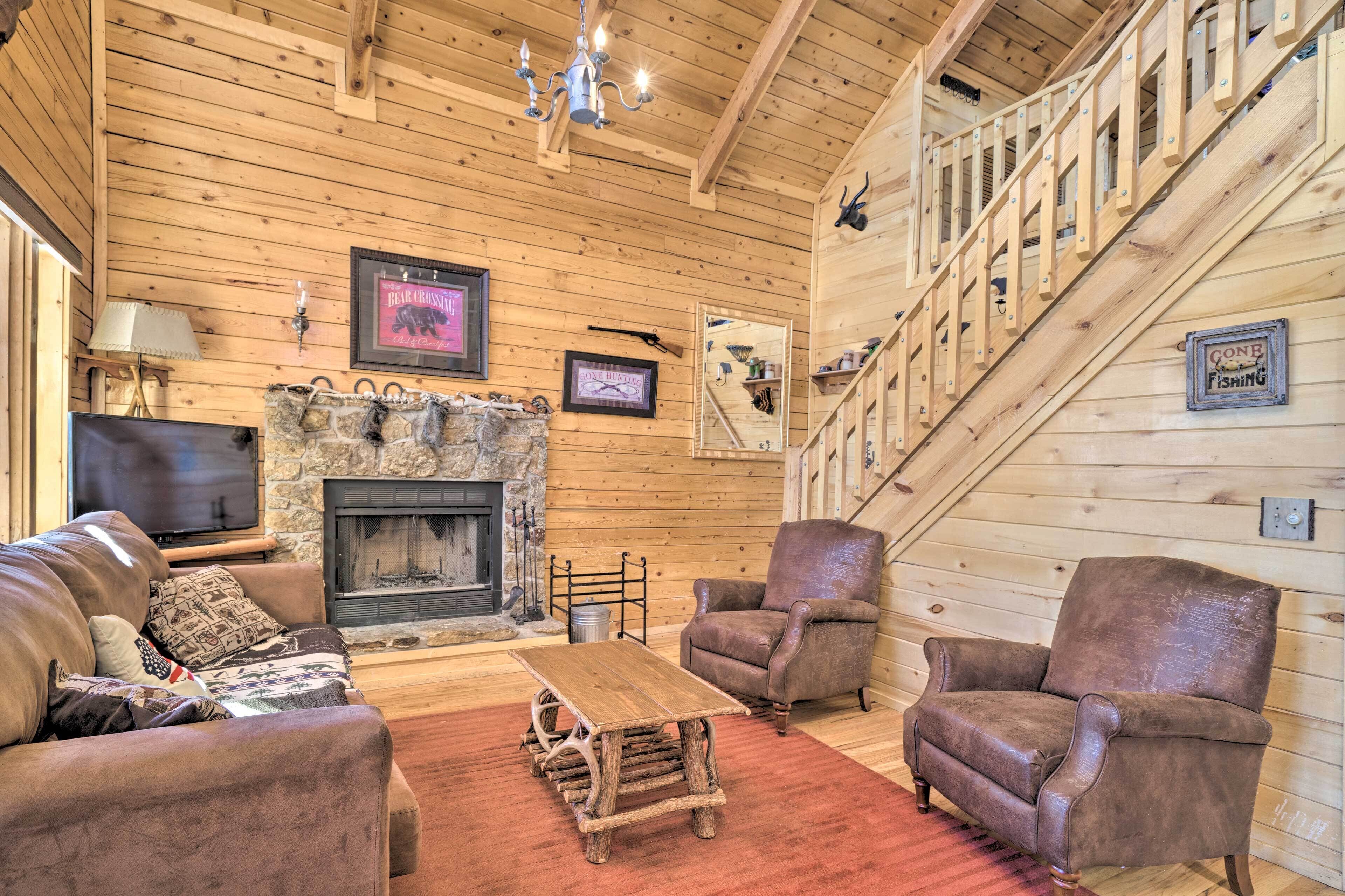 A cozy log cabin interior with a stone fireplace, leather furniture, and a staircase.
