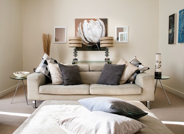 A living room with a beige sofa, multiple pillows, a glass coffee table, a vase with dried plants, and a painting on the wall.