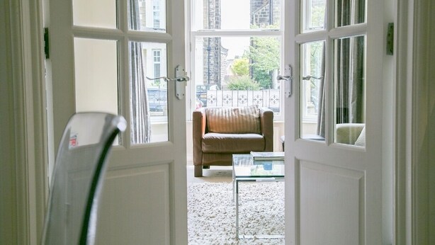 Een zicht door een deuropening naar een kamer met een bruine leren bank, een glazen tafel en een wit tapijt.