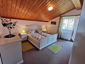 A bedroom with a wooden ceiling, a bed with floral bedding, bedside lamps, a vase with flowers, and a window with curtains.