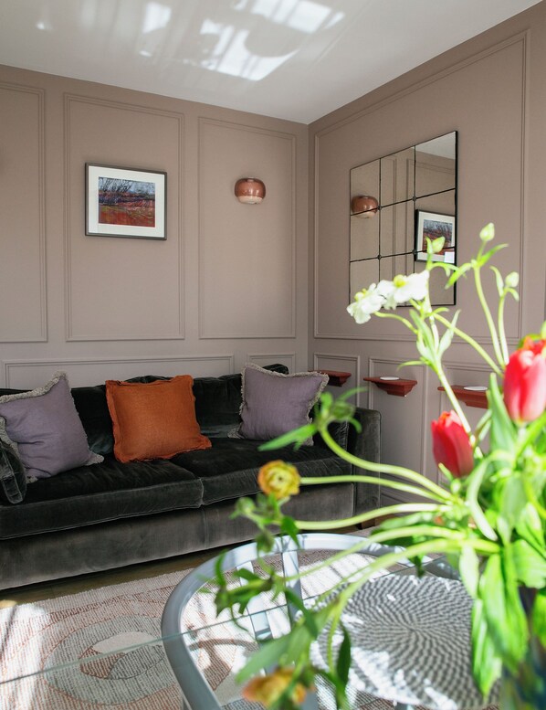 A living room with a dark sofa, decorative pillows, a round glass table with a vase of flowers, and a large mirror on the wall.