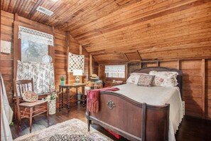 A wooden cabin bedroom with a bed, a chair, a table, and a window with curtains.