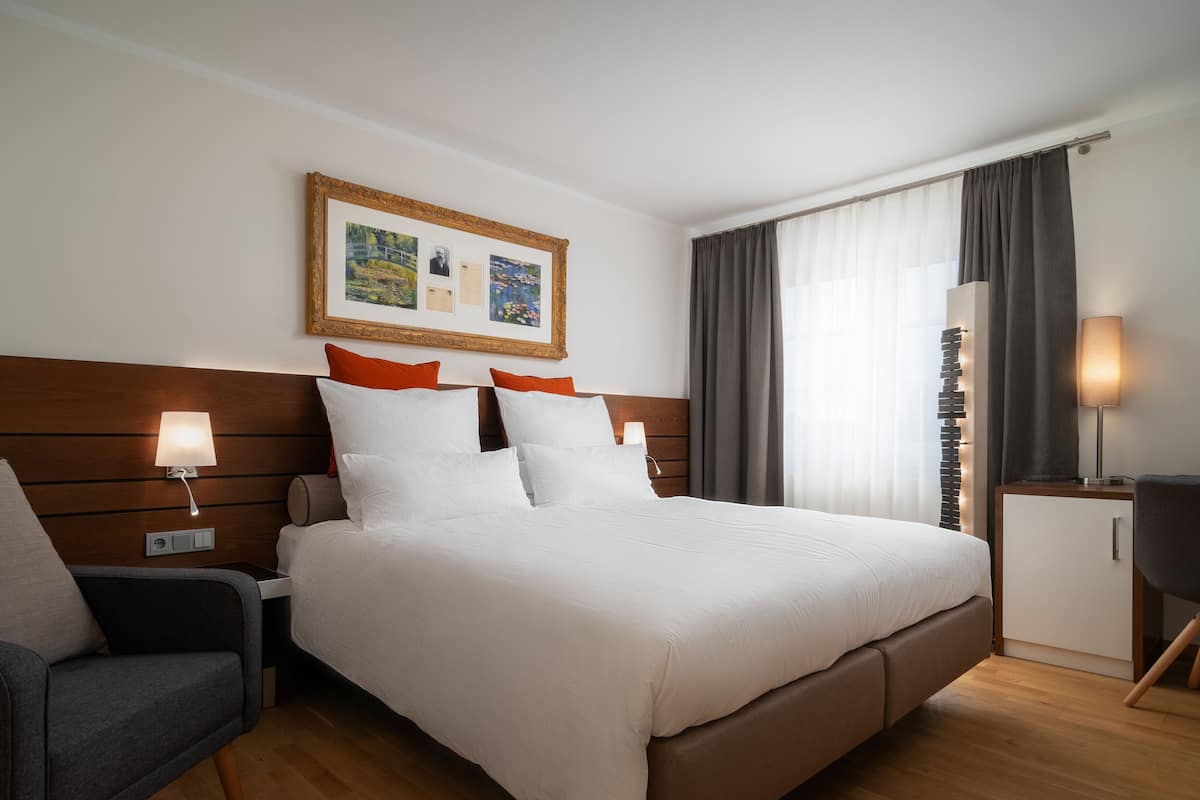 Quarto de hotel com uma cama grande, mesa de cabeceira com abajur, uma cadeira, escrivaninha com abajur e um quadro na parede.