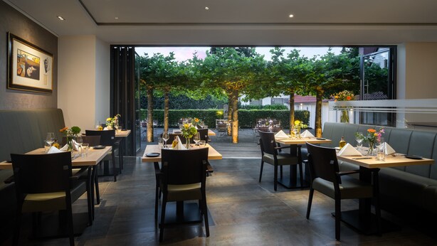 An indoor dining area with tables and chairs, a sofa, and a large window overlooking a garden. - Hotel zur Post Riesenbeck (Hoerstel)