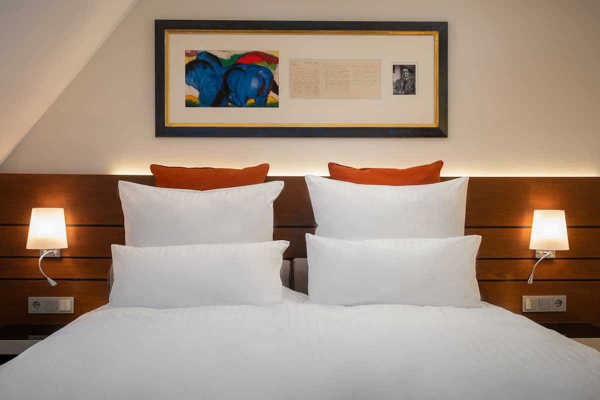 Quarto de hotel com uma cama grande, duas almofadas brancas, duas almofadas laranjas, uma obra de arte emoldurada acima da cama e duas luminárias de cabeceira.