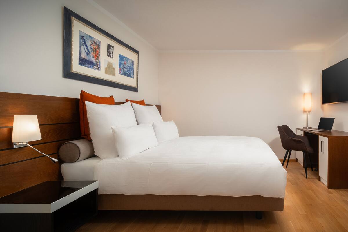 Quarto de hotel com cama, mesa de cabeceira, abajur, escrivaninha, cadeira e televisão de tela plana fixada na parede.