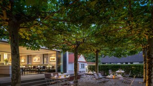 An outdoor dining area with tables and chairs, surrounded by trees, and a building with a red roof in the background. - Hotel zur Post Riesenbeck (Hoerstel)