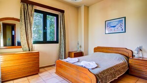 A bedroom with a wooden bed, a dresser, a nightstand, a window with curtains, a wall-mounted lamp, and a framed picture. - Peaceful two storey villa with traditional aesthetics (Heraklion)