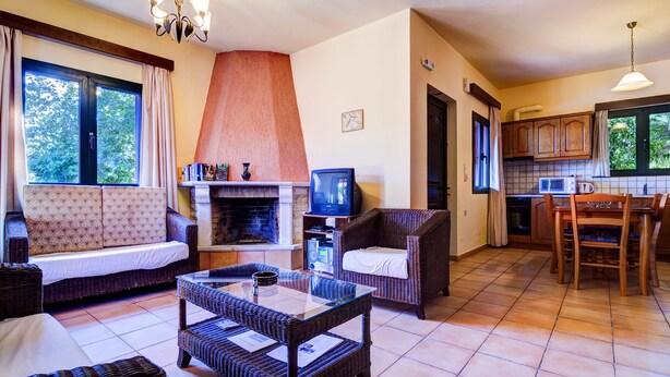 A living room with a fireplace, sofa, and wicker furniture. There is a television and a kitchen area visible. - Peaceful two storey villa with traditional aesthetics (Heraklion)
