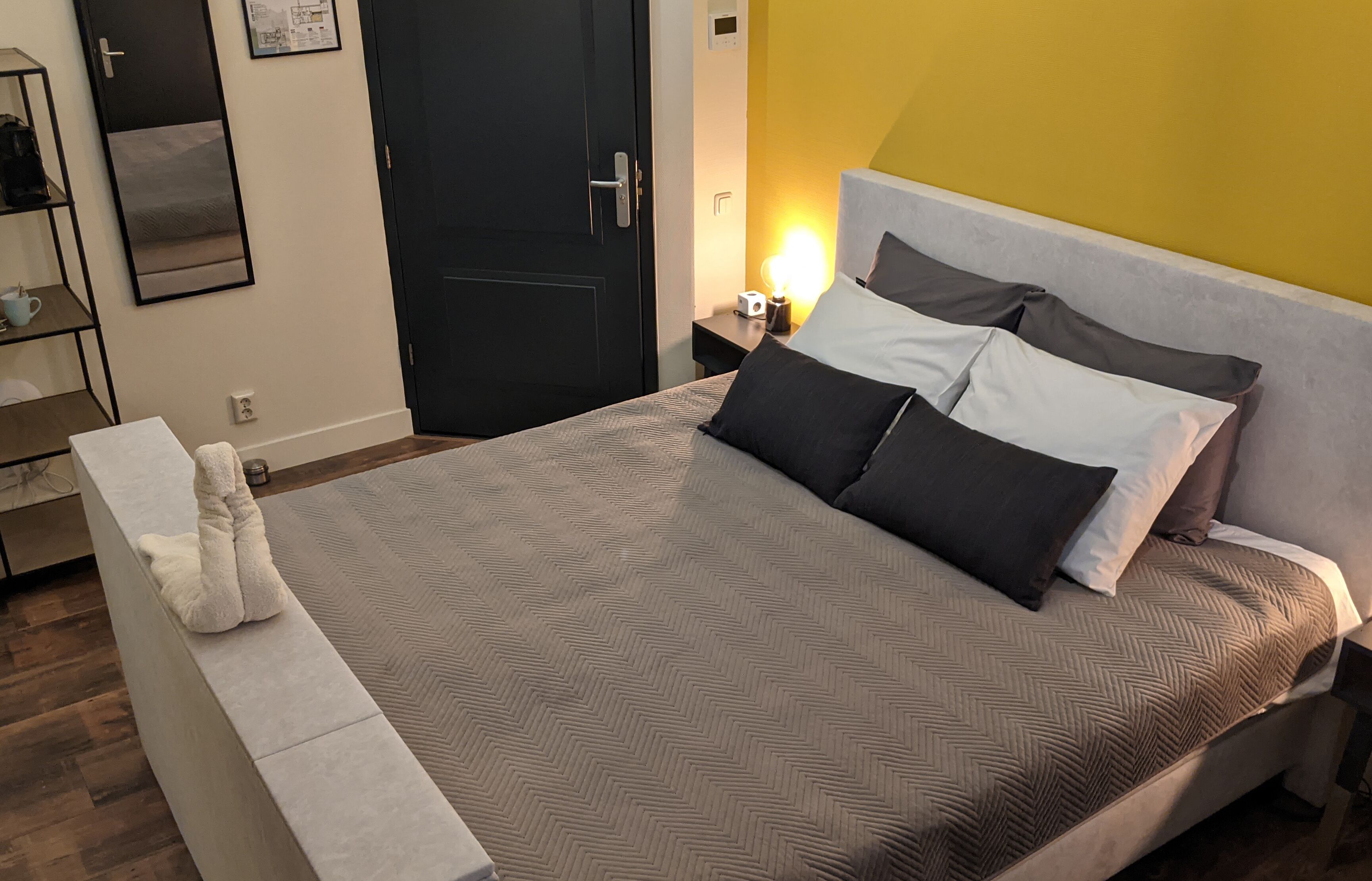 Een net opgemaakt bed met een grijs-wit beddegoed, een witte nachtkastje, een zwarte deur en een gele muur.