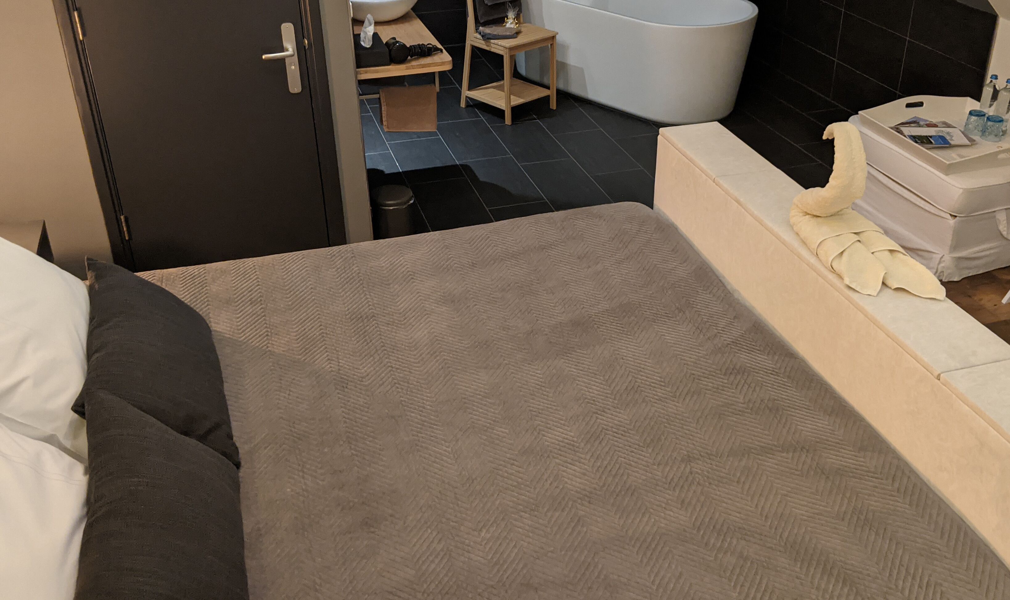 A bedroom with a bed, a bedside table, a small wooden stool, a white bathtub, and a bathroom counter with toiletries.