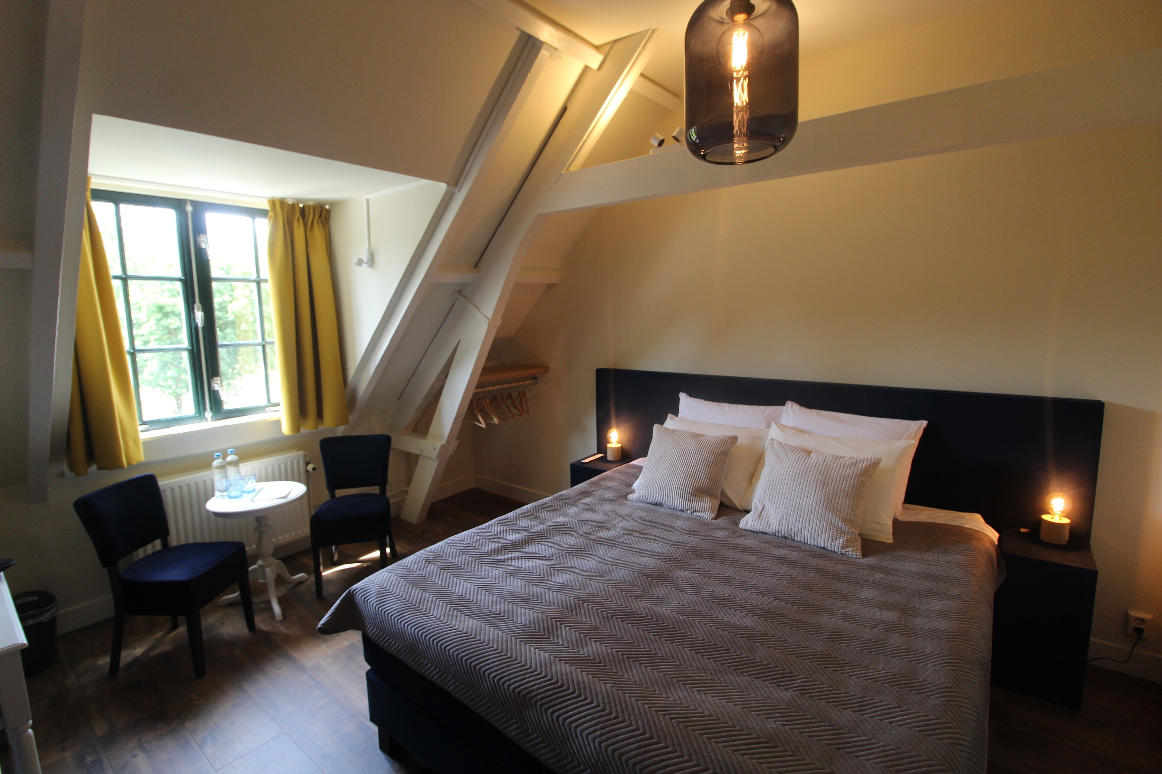 Een slaapkamer met een groot bed, een bureau met stoelen, een raam met gordijnen en een schuine kap met een lamp.