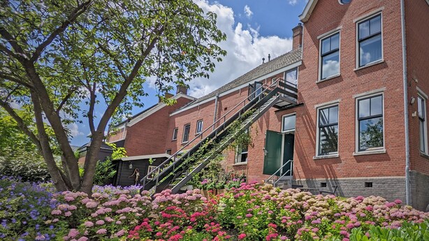 A red brick building with a staircase leading to the entrance, surrounded by a garden with pink and purple flowers. - Polderhuis Bed & Breakfast (Bergschenhoek)
